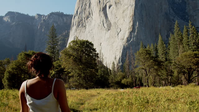 Young attractive woman walking in meadow looking up at El Capitan rock face in Yosemite National Park