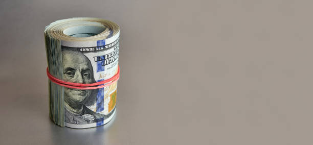 istock Many hundred dollar bills rolled up with a red rubber band on a colored background. 1671176221