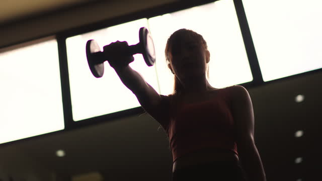 Low angle view of beautiful Asian lady doing a dumbbell front raise exercise inside the gym during the evening