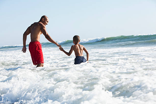 Father and son playing in surf Black father (30s) and son (6 years) at the beach, playing in the surf. waist deep in water stock pictures, royalty-free photos & images