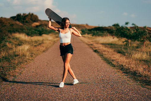 Beautiful young woman listening to music through headphones holding a skateboard on the road during a sunny day