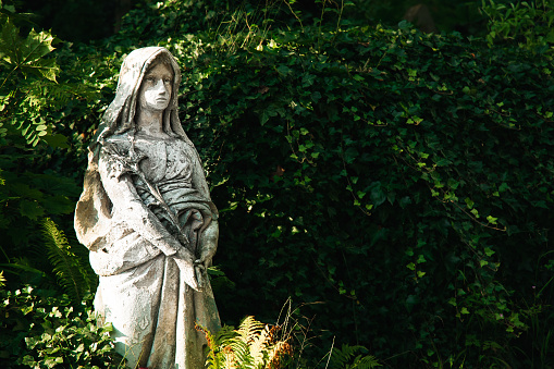 Religious sculpture of nun stone material figure in parkland environment with with green blurry foliage of bush