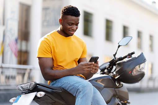 Calm african american man using mobile phone while relaxing at the street after motorbike riding. People lifestyle concept
