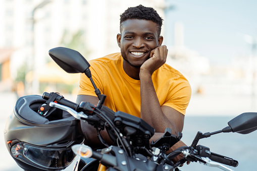 Smiling confident african american man, biker holding helmet and looking at camera near sport motorcycle. Handsome model wearing orange t shirt posing for pictures