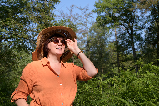 A woman in a shady green park. Orange dress, straw hat, sunglasses. Place for text