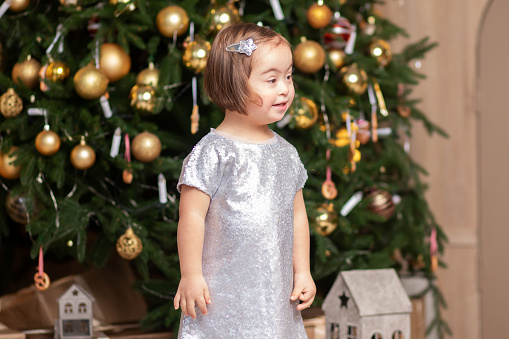 happy girl with down syndrome in a festive dress is standing near the Christmas tree, smiling