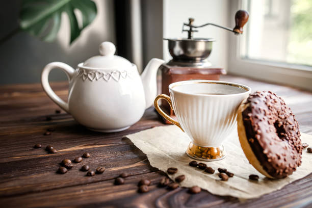 white kettle and cup on table. Breakfast by window. Chocolate donat next to scattered coffee beans.