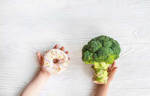 baby's hands holding sweet Donat and broccoli. Choosing choosing between Vegetables or junk food for toddlers. Healthy clean detox eating concept. Children's dietary nutrition . Copy space