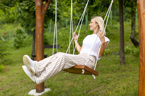 Beautiful woman looks very happy and cheerful while swinging in backyard on countryside. It is a springtime and sunny day.