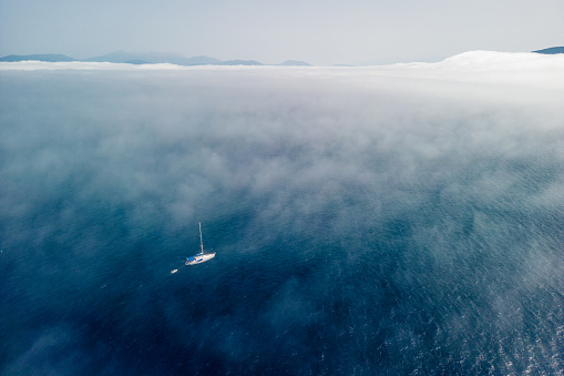 Sailboat sailing through fog over the Ionian Sea. Greek island rises above the low fog, and the sky is clear and blue. Aerial view from above is made with a drone during the sailing.