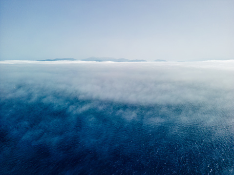 Beautiful landscape with fog over the Ionian Sea. Greek island rises above the low fog, and the sky is clear and blue. Aerial view from above is made with a drone during the sailing.