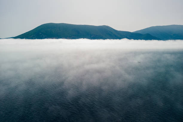 Sailboat sailing through fog over the Ionian Sea. Greek island rises above the low fog, and the sky is clear and blue. Aerial view from above is made with a drone during the sailing.
