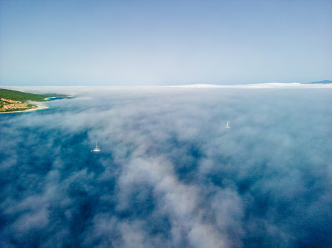 Sailboat sailing through fog over the Ionian Sea. Greek island rises above the low fog, and the sky is clear and blue. Aerial view from above is made with a drone during the sailing.