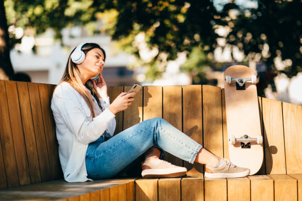 Young woman sitting on a bench in the city and listens to music via smartphone and headphones during a sunny day stock photo