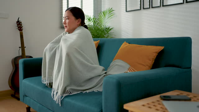 Asian woman feel cold, wrapped in blanket sitting on sofa in living room at home.