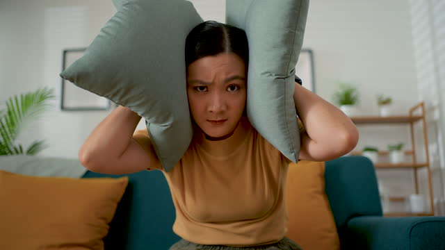 Asian woman feel annoyed with noise covering ears by pillows sitting at home.