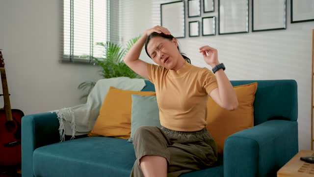 Asian woman feels pain from injury neck and shoulder sitting on sofa in living room at home.
