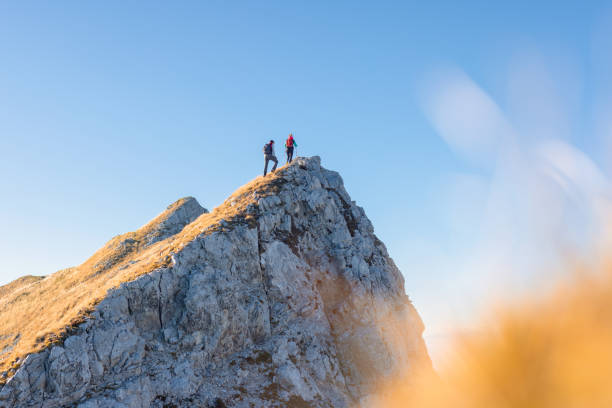 Two hikers walking along a mountain ridge route, the sun on the blue sky Spectacular view of two hikers walking along the mountain ridge route with the sun on the clear blue sky, aerial shot. Inspiration, nature, and mountaineering concepts. mountain peak stock pictures, royalty-free photos & images