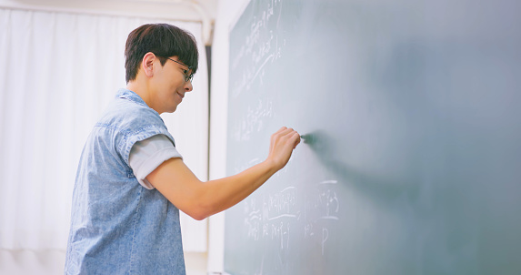 asian male teacher or college student is writing on blackboard during class in classroom at university