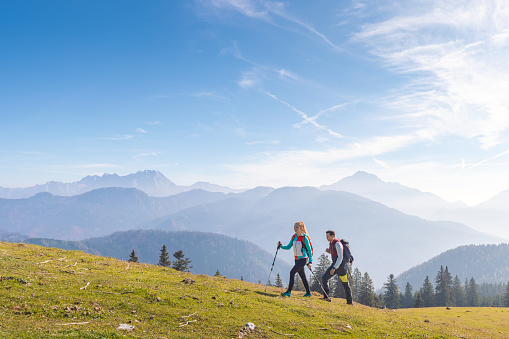 Two hikers, a man and a woman, walking over a grassy slope with a scenic view of misty mountains under the morning autumn sun in the background