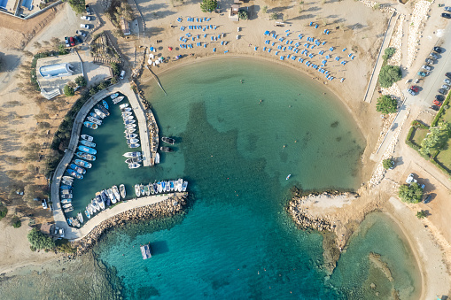 Aerial drone view of fishing harbour and sandy beach. Agia Triada people swimming and fishing boats moored at the harbor. Protaras Paralimni Cyprus