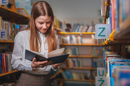 Young male student concentrated reading a book in the library while standing in back of library alone.