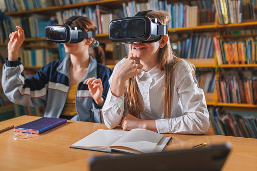 Two female students studying with VR headsets in the library, sitting at a desk with a laptop and notebooks. Concept of modern library and education.
