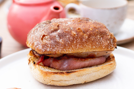 Bacon sausage sandwich on a crispy roll with butter and tea pot in background. Close up style