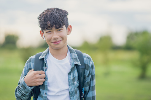 A young boy of Asian decent, stands outside as he poses for a school portrait.  He is dressed casually and smiling as he holds his backpack over his shoulders.