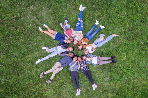 A large group of ethnically diverse teens, lay in the grass in a circle as they pose for a portrait.  They are each dressed casually and are smiling in this aerial view.