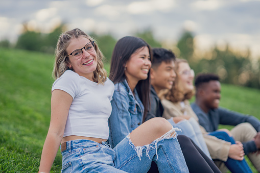 A small group of teens sit on a grassy hill side-by-side as they hang out together.  They are each dressed casually as they enjoy the fresh air and each others comapny.