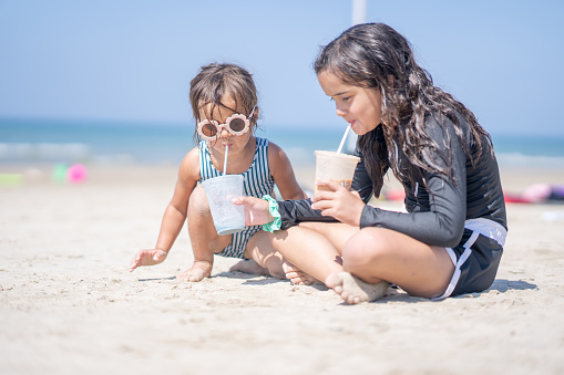 Two sisters sit on the beach together as they cool off with a drink.  They are both wearing swim suits and are focused on sipping their drinks.