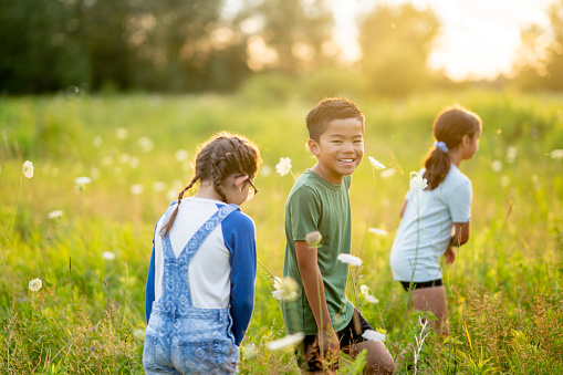 A small group of school aged children are seen walking through a field of tall grass as they take a walk and explore together.  They are each dressed casually and are smiling as they enjoy the warm summer air.