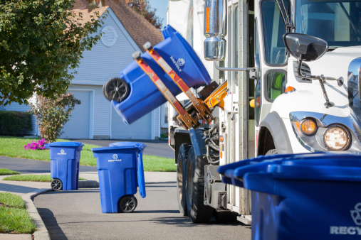 A recycling truck in a residential neighborhood is shown using it's mechanical arm to lift up a blue container containing recyclable materials. Other containers can be seen along the curbside. Canon 5D MarkII. 