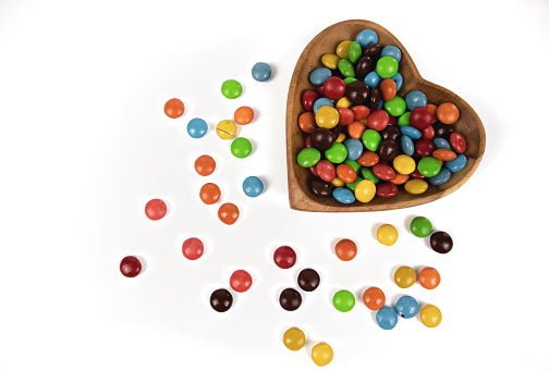 pile of colorful chocolate candies in heart shaped wooden bowl isolated on white background, top view