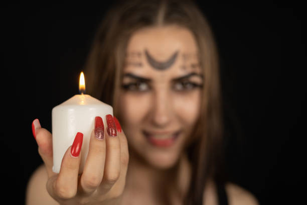 In the evening of Halloween, the witch, smiling cutely, holds out a burning candle to the camera. On a black background, a woman in the form of a witch with magic symbols on her face holds a candle. In the evening of Halloween, the witch, smiling cutely, holds out a burning candle to the camera. On a black background, a woman in the form of a witch with magic symbols on her face holds a candle.. cutely stock pictures, royalty-free photos & images