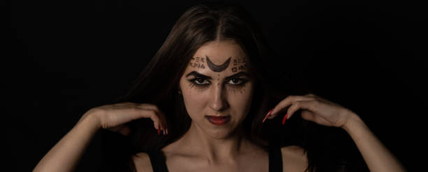 on halloween night, a witch with red nails straightens her long hair with her hands and stares piercingly into the camera. porter. a female witch with magical symbols on her face is looking at you. - witch voodoo smiling bizarre imagens e fotografias de stock