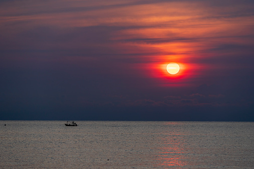 Distant photo of a small boat and an identifiable fisherman in silhouette. Sailing on the sea near the horizon There is a rising or setting sun,depending on the viewer's interpretation and application