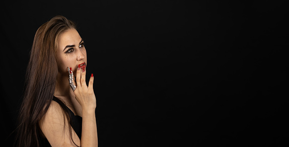 Sexy gothic witch for halloween on a black background looks towards the empty space for insertion. A woman in the form of a witch holds her hand to her face and looks away. Banner, flyer..