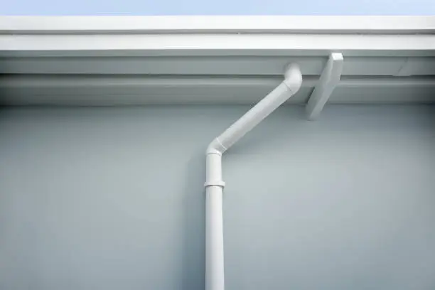 Rain gutter install on steel structure, connect to pvc downpipe or downspout, elbow at eaves, exterior house building. Also called guttering or eavestrough for water drainage system. Look new clean.