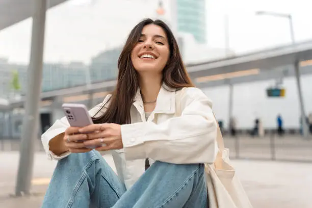 Photo of Waiting for the bus and for love. Bright cheerful young woman of Turkish mixed race in white casual clothes with a charming smile holding a phone in the street at public transportation stop