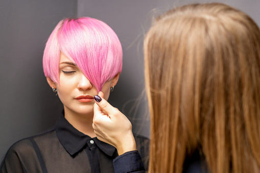 Hairdresser with hands is checking out and fixing the short pink hairstyle of the young white woman in a hair salon