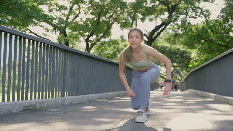 Young Asian woman ties her shoelaces and prepares for a jog on an elevated walkway flanked by trees in a sunny park.