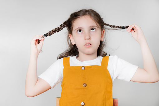 Portrait of cherubic blue-eyed little girl titivating touching short curly fair hair, wearing pink jumpsuit, bracelets, looking at camera, posing on blue background. Childhood, copy space, studio.