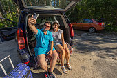 A couple of travelers stopped in a parking lot in the woods and, sitting in the trunk of a car, took a selfie with their phone. A man and a woman drive a car and go to relax and take a selfie.