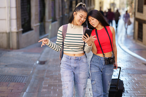 Young positive Asian woman with backpack wearing striped sweater, and jeans showing smartphone to friend walking with photo camera and suitcase pointing at street during trip through Granada