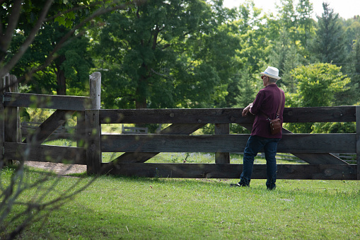 A man is standing at a fence. It is a rural location. The fence is wooden, with 3 large  horizontal panels. Trees are in the background