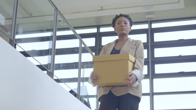 African American woman hold box and walk down from stair to lower level or floor look like she quit or lose job and take her things to go out from office.