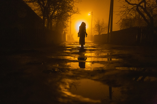 Lonely woman walking in fog away road. Rural landscape with road in morning mist. Warm autumn colors. Dark mysterious background