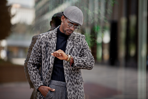 Handsome black man in stylish clothing looking at wristwatch while standing near glass wall in city. He is wearing trendy cap, coat and glasses.